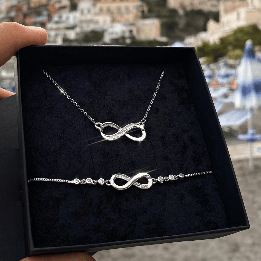 Silver infinity necklace and bracelet set in a black box with a blurred outdoor background.
