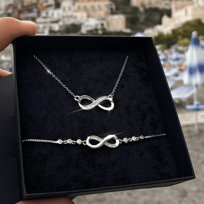 Silver infinity necklace and bracelet set in a black box with a blurred outdoor background.